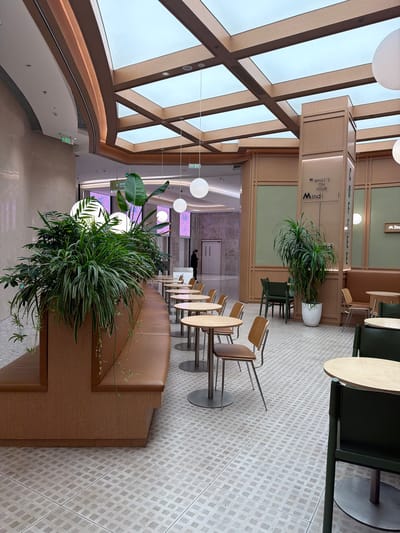 The image depicts an interior view of a modern commercial space, likely a café or lounge, located in the Jianye District of China. The space features a high ceiling with a geometric, skylight structure supported by wooden beams.

The foreground is dominated by a large, lush planter filled with various green plants, situated on top of a cushioned bench. Adjacent to the bench are several wooden chairs with metal legs arranged at small, round tables.

Beyond the seating area, there is a corridor leading further into the building, with a person visible in the distance. The space is well-lit by overhead lighting and natural light filtering through the skylight. The floors are covered in light-colored tiles arranged in a grid pattern.
A sign reading "What's on your Mind" is visible near another plant.
The atmosphere appears modern and inviting.