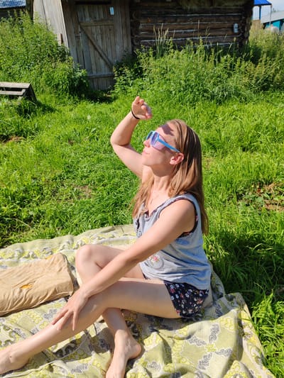 In a rural outdoor setting, likely in or around the city of Chukhlom, Russia, a woman sits on a blanket on a grassy lawn. She is positioned in a relaxed, seated pose with her legs crossed and her left arm raised, hand shading her eyes as she looks upwards. She is wearing a gray sleeveless top with a floral pattern on the neckline and dark shorts with a floral print. Blue-rimmed sunglasses rest on her nose. Beside her on the blanket is a light brown pillow. Behind the woman, a wooden structure, possibly a shed or outbuilding, stands amidst tall green grass and vegetation. The overall scene suggests a sunny day, possibly for leisure or relaxation outdoors.