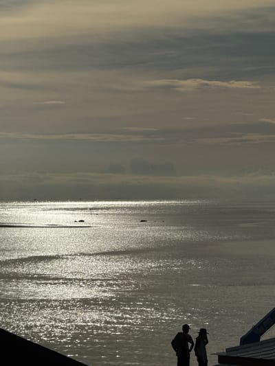 The image shows a silhouetted view of two people standing on a dark structure overlooking a body of water. The water is illuminated by the sun, creating a shimmering effect. In the distance, there are several small boats and what appear to be rocks or small islands. The sky is cloudy, with layers of light and dark clouds. The scene is likely taken at sunset or sunrise, given the lighting conditions. The location is identified as Ko Phangan, Thailand.