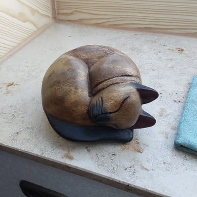The image shows a wooden carving of a sleeping cat. The cat is curled up in a ball with its head tucked under its body. The carving is made of dark wood and is painted brown. The cat is resting on a white surface with a blue cloth to the right. The surface is covered in white specks. This location is on the edge of a building in Garbsen, Germany. The building is made of white brick and has a wooden trim. The building is located on a street with a sidewalk in front of it.