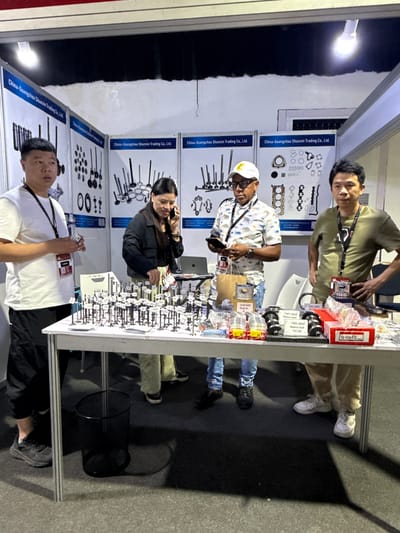 The image shows a group of people at a trade show booth. There are four people visible, likely employees or representatives of the company. They are standing behind a table displaying various automotive parts. The backdrop of the booth features the name "China-Guangzhou Shunxin Trading Co., Ltd." along with visual representations of engine components. Various parts are on display, with some labeled as piston kits and balance shafts. One person is using a phone, while others appear to be engaging with potential customers or handling products. The booth is well-lit, suggesting an indoor trade show environment. The location in Itirin, Nigeria is not evident in the image.