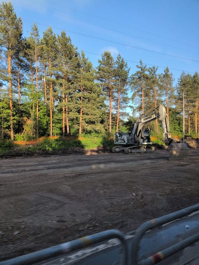 The image depicts an excavator, identified by the "LIEBHERR" branding on its boom, situated on a cleared construction site. The site features disturbed earth and tracks from heavy machinery. Behind the construction area, a dense line of tall pine trees forms a natural barrier, with some younger green foliage and an orange safety fence visible among them. The sky is clear and blue, with faint power lines stretched across the upper portion of the frame. The overall scene suggests ongoing construction work near a wooded area, likely in a peripheral or developing part of Vantaa, Finland, given the presence of pine trees typical of the region. There are no people visible in the image, and the excavator appears to be stationary. The foreground is blurred, indicating the photo was taken from a moving vehicle.