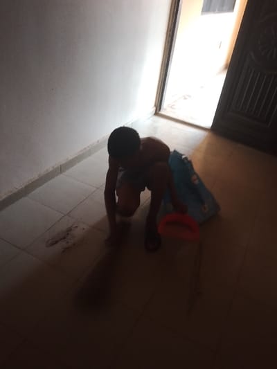 The scene depicts an interior space, identified as being in Ufuma, Nigeria. A single individual, appearing to be a young male, is crouched low on a light-colored tiled floor. He is shirtless, wears dark shorts and dark flip-flops, and is seen from a rear-left perspective. His left hand is positioned near a dark, irregular stain or residue, approximately 15-20 cm in length, on the floor. An orange, U-shaped object, possibly a small bucket or scoop, rests on the floor to his right. Behind him is a crumpled light blue object, likely a bag or cloth, displaying white circular patterns. The room features light-colored walls with a dark skirting board. To the right, an open dark-colored door frame reveals a brightly lit area beyond. A portion of a dark door with an embossed or carved design is visible on the far right. The immediate foreground and the individual are in shadow, while the area beyond the doorway is brightly illuminated.