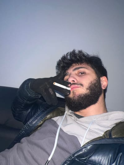 A man with dark hair and a beard is pictured at an angle, gazing at the camera. He wears black gloves and holds a lit cigarette near his mouth. He is clad in a grey and white hoodie, partially obscured by a puffy black jacket. The background is a plain, light grey wall. The lighting is dim, casting shadows and softening the details of the image.
