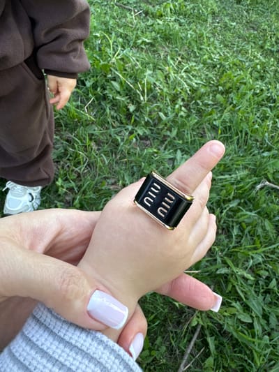 A close-up, overhead shot shows an adult hand holding a child's hand. The adult is wearing a gray, ribbed sweater. The adult's fingernails are manicured in a French style. The child is wearing a brown hoodie. The child's index finger has a large, rectangular black ring with the word "MIU MIU" printed on it twice. Both are outside on green grass. A child's white sneaker is partly visible on the grass. The photo was likely taken in a park or grassy area. This image does not show the location of Odintsovo, Russia.