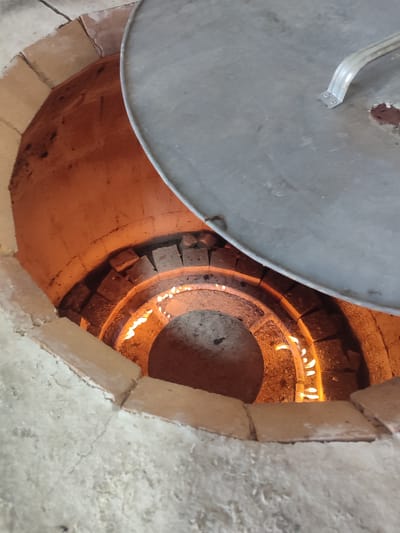 An overhead shot shows the interior of a clay tandoor oven with a large, circular metal lid partially covering the opening. The inside of the oven glows with orange heat from a visible fire burning at the bottom. The walls of the oven are constructed from rough, beige-colored clay and appear to be made of stacked bricks in some areas. The lid is metallic and has a simple, curved handle attached with small screws. The surrounding area outside the oven appears to be a rough, concrete-like surface. The image was taken in Жаңа Қуат, Kazakhstan.
