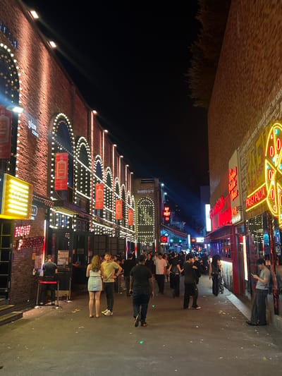 The image shows a bustling street scene in Seoul, South Korea, at night. A crowd of people is walking along the sidewalk past a row of buildings with brightly lit facades. The buildings are decorated with strings of lights and neon signs. The signs include the name "Peggy Pizza" and "Glam Lounge". The lighting is vibrant, creating a lively atmosphere. The sky is dark, and there is sparse greenery above the structures.