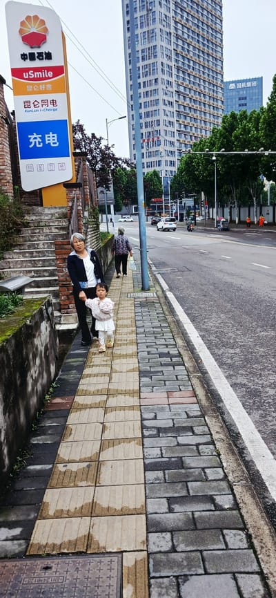 Here is a detailed description of the image:

**Setting:** The image appears to be taken on a sidewalk next to a street in the Jiulongpo District of China, judging by the presence of Chinese characters on the sign and the overall urban environment. Tall buildings are visible in the background, indicating a densely populated area.

**People:** The image captures several individuals. A woman is seen holding a young child's hand. Another elderly woman is walking alone nearby.

**Objects:** Prominently featured in the foreground is a sign with Chinese characters and the term "uSmile," "KunLun i-charge" and the logo of China National Petroleum Corporation (CNPC). The sign appears to be advertising charging services. The sidewalk is constructed of interlocking paving stones, some of which appear wet, which is likely due to rain. There is also a manhole cover on the sidewalk. Vehicles are on the road in the background.

**Actions and Interactions:** The woman is holding hands with the child, suggesting she is helping the child walk. The elderly woman is walking independently. Cars are moving along the street.

**Environment:** The environment appears to be urban, with tall buildings lining the street. The presence of trees indicates there might be landscaping along the road. The image suggests a cloudy day, as the sky is overcast and the sidewalk looks wet.
