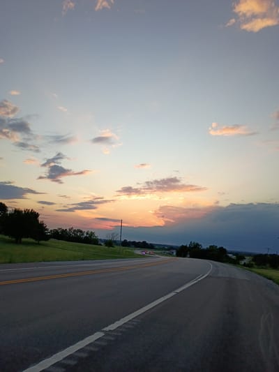 A low-angle, wide shot captures a paved road curving to the right under a twilight sky. The road, divided by double yellow lines and a dashed white line on the right, appears to be a two-lane highway. Flanking the road on the left is a grassy embankment dotted with trees. On the right, the road meets a field and more trees. In the distance, lights from a few vehicles are visible on the highway, and a utility pole stands near the roadside. The sky is a blend of pale blue and soft orange, with scattered clouds tinged pink and yellow by the setting sun. The overall scene suggests a rural or suburban setting, possibly outside of Lancaster, United States, given the landscape and road type.