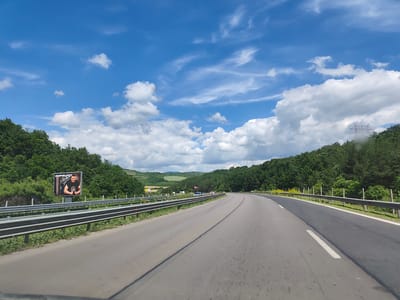 This is a forward-facing, eye-level, medium-distance shot of a highway in Potop, Bulgaria, on a sunny day with a blue sky and scattered clouds. The highway curves to the right, with a clear lane division. On the left side of the highway, there is a metal guardrail, and beyond that, a billboard featuring a man with his thumbs up, wearing a black t-shirt. The background consists of a dense forest on either side of the road, with rolling hills and some power lines visible in the distance. Several vehicles are present on the highway ahead.