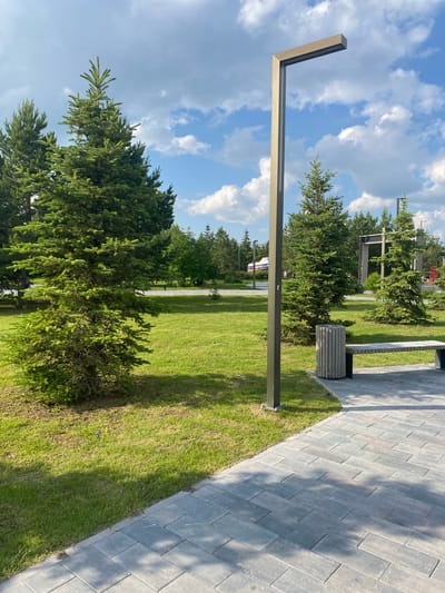 This outdoor image depicts a well-maintained public park or green space in Tyumen, Russia, specifically identified by a large, blue and white boat-like installation in the distant left background displaying the text "ВЕЛИКАЯ ОБЬ" (Great Ob).

In the foreground and mid-ground, a light gray paved walkway, constructed from rectangular pavers laid in a staggered pattern, extends from the bottom right towards the center. The walkway is bordered on the left by lush green grass. A tall, dark gray, L-shaped modern street light pole stands prominently on the right side of the path, featuring an integrated LED light fixture at its horizontal top section. To the right of the light pole, a contemporary park bench with a light-colored wooden slatted seat and a dark metal frame is partially visible, next to a matching cylindrical wooden slatted trash receptacle.

Numerous evergreen trees, primarily firs or spruces, are dispersed throughout the grassy areas, with a large, vibrant green fir tree positioned prominently in the mid-left. In the distant background, beyond the trees, a modern, open-sided structure with a dark frame is partially visible. Two indistinct human figures appear small in the far distance, near the "ВЕЛИКАЯ ОБЬ" installation. The scene is illuminated by natural sunlight, casting distinct shadows from the trees and light pole onto the grass and pavement. The sky above is clear blue with scattered white cumulus clouds.