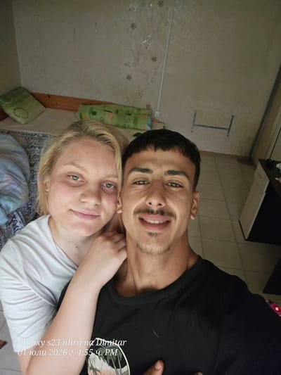 This media file features a close-up, selfie-style photo of a young couple. The man, with dark hair and a mustache, is positioned behind and to the side of the woman, with his head resting on her shoulder. He is smiling, with his eyes looking towards the camera. The woman, with blonde hair, is facing the camera and also smiling. They appear to be indoors, in a casual setting, possibly a bedroom or living area, given the presence of a bed with patterned bedding and a pillow. A green patterned fabric hangs on the wall behind them, and a wooden beam is visible. The lighting suggests daytime. The image contains visible text which appears to be a timestamp and camera model: "Galaxy s23 ultra na Dimitar 01 юли 2026 г. 4:55 ч. РМ". The overall mood is affectionate and relaxed.