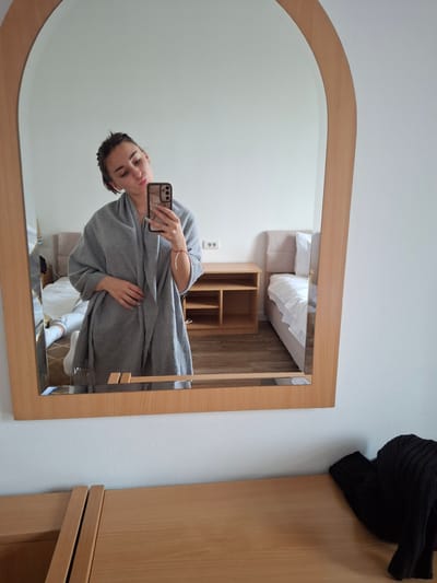 The image presents a candid scene within a bedroom in the city of Vozdovac Urban Municipality, Serbia. A young adult female is visible in the reflection of a large, vertically oriented mirror with a light-colored wooden frame and an arched top. She is taking a selfie using a dark-colored smartphone, held in her right hand, and is pouting her lips. Her dark hair is pulled back, and she is wrapped in a grey shawl or blanket. A necklace is visible around her neck, and a red and white cord bracelet is on her left wrist.

The reflected background reveals a sparse room with white walls and a light wood-patterned floor. To the right, a bed with a light beige upholstered headboard and white bedding is visible, alongside a matching light wood nightstand featuring open shelves. To the left of the primary subject, the lower legs of another individual, wearing grey sweatpants and white socks, are visible lying on what appears to be a separate bed or surface. A double electrical outlet is mounted on the white wall in the background. In the foreground, below the mirror, a light-colored wooden dresser or table surface is present, upon which a folded black ribbed knit item, possibly a sweater or beanie, rests on the far right.