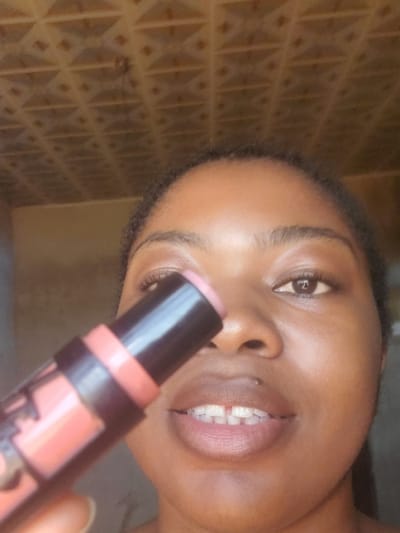 The scene is depicted in Kakau, Nigeria. A close-up shot features an individual, likely female, with dark skin and dark, short, curly hair framing the forehead. The individual's dark brown eyes are open, with visible eyelashes, and their gaze is directed towards the viewer. A small, silver piercing is visible on the right side of the upper lip. The mouth is slightly open, revealing white teeth with a distinct gap between the two central incisors. The individual holds a black and coral-pink makeup stick in the foreground, with the light pink product tip extended. The stick is positioned diagonally across the lower-left portion of the face, obscuring part of the lower jaw and neck. The background consists of a light brown, patterned ceiling with a grid of raised squares or diamonds in the upper half of the frame. Below the ceiling, a plain, light brown wall is visible, out of focus.