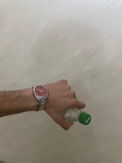 A person's forearm and hand, adorned with a watch, are centered against a light-colored, textured wall. The hand holds a small, clear plastic bottle with a green screw cap. The background wall exhibits subtle brush strokes or texture, suggesting an indoor setting in Ihtiman, Bulgaria.

The watch on the person's left wrist is silver-toned with a metal bracelet and features a prominent red dial. The dial displays white Roman numeral hour markers, a date window at the 3 o'clock position, and white hour, minute, and second hands. A fluted bezel surrounds the watch face. The brand "CASIO" is faintly discernible on the upper part of the dial, and "WR 50M" (Water Resistant 50 meters) is partially legible below the center.

The bottle, held by the fingertips, contains a clear liquid. The green cap clearly shows the word "Sprite" in white text, indicating its brand. The arm is visibly hairy. The image captures a casual, everyday moment.