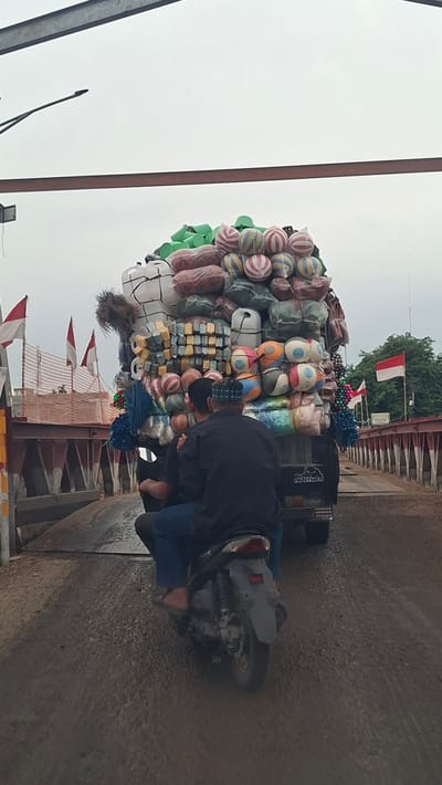 In Kulukuta, Indonesia, a heavily overloaded truck is captured on camera, its cargo a mountain of various goods, predominantly brightly colored inflatable balls and large white plastic jugs. The truck is partially obscured by a motorcycle carrying two individuals, their backs to the camera. The rider wears a black long-sleeved shirt and blue pants, while the passenger has on a dark shirt and a patterned skullcap. They are riding on a wet, dusty road, suggesting recent rain. The scene is set on a bridge, indicated by the red metal railings on the right side of the frame. Indonesian flags are visible on the bridge and on the truck, adding a nationalistic element to the otherwise bustling and chaotic transportation scene. The sky is overcast, contributing to the muted lighting of the photograph.