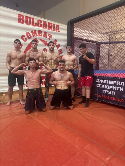 Seven young men, appearing to be athletes, are posing in a combat sports arena. The backdrop features a large banner with red lettering that reads "BULGARIA COMBAT SAM". To the right, a fenced-in cage, commonly used in MMA or wrestling, is visible, with a blue mat below it. A black banner with white and red text, including Cyrillic script and a phone number, is attached to the cage. The floor of the arena is an orange mat. The men are shirtless or wearing athletic shorts and t-shirts, and some are flexing their muscles. One man in the front row is kneeling and flexing, while another is giving a peace sign. The scene suggests a training session or a team photo at a combat sports facility in Gorna Oryahovitsa, Bulgaria, likely associated with the "Bulgaria Combat Sam" organization.