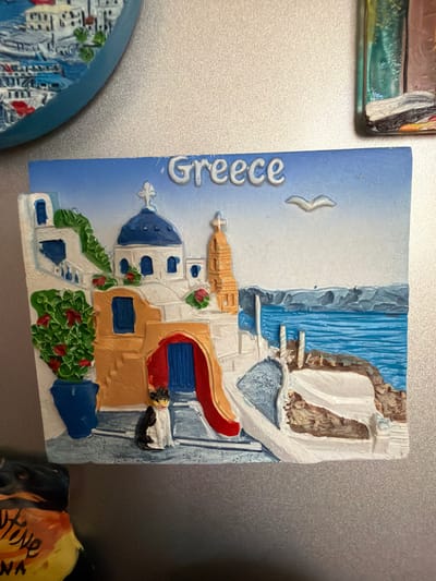 The image is a close-up photograph of a souvenir magnet depicting a scene from Greece. The magnet features white buildings with blue domes and accents, characteristic of Greek architecture. There is a prominent blue ocean in the background with distant islands. A small black and white cat is positioned on the cobblestone path in the foreground. The word "Greece" is written in raised, white lettering at the top of the magnet. The magnet is attached to a metallic surface, likely a refrigerator. The photograph does not depict Pazardzhik, Bulgaria.
