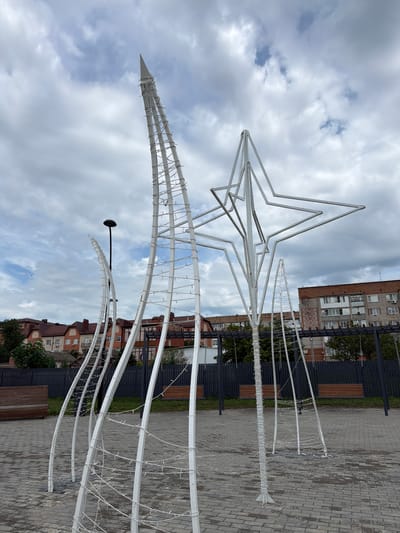 The image depicts an outdoor scene in Leningradskaya, Russia, featuring a large, white, abstract metal sculpture resembling a star and a towering spike. The sculpture is adorned with string lights. In the background, residential buildings and trees are visible. The ground is paved with gray bricks, and there are benches and a lamppost. The sky is cloudy.