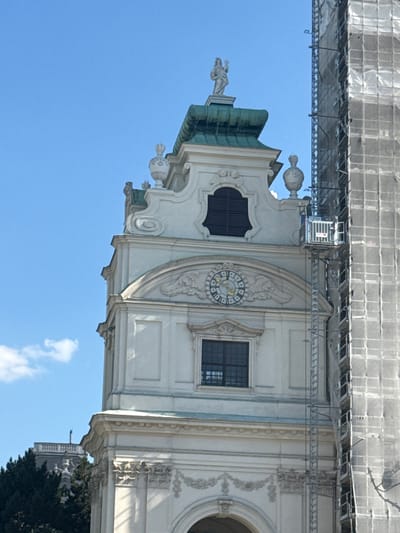 The image shows a close-up of a tall, ornate white building with a green roof, set against a clear blue sky with a few wispy clouds. The building features classical architectural elements, including decorative moldings, arched windows, and a statue atop the roof. A clock face is visible on the facade, with Roman numerals. To the right of this building, a tall structure covered in scaffolding and white netting is partially visible, suggesting ongoing construction or renovation work. In the background, another building with a distinctive roofline and a chimney can be seen peeking out from behind trees. The scene is likely a historical district in Vienna, Austria, given the architectural style.