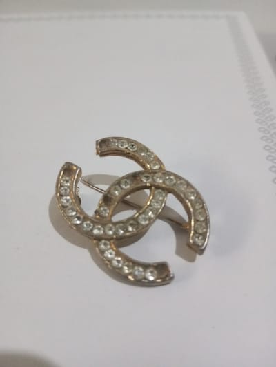 The scene is an overhead, slightly angled close-up of a metallic brooch resting on a plain white surface in Ciherang, Indonesia. The brooch features two interlocking 'C' shapes, creating a double C logo. It is crafted from a light-colored metal, possibly a silver or gold tone, which exhibits visible wear, discoloration, and minor pitting in several areas. Each 'C' is encrusted with multiple small, clear, round faceted stones, consistent with rhinestones or cubic zirconia, approximately 3-4mm in diameter. Some stones appear slightly dull or scratched. A standard pin mechanism, consisting of a straight pin and a C-clasp, is visible on the rear of the brooch. The brooch casts a soft shadow to its lower right. The white surface extends across the frame, with faint, parallel embossed lines or a texture visible on the upper right quadrant, possibly indicating paper or a textured table. No individuals or discernible actions are present within the frame. The environment appears to be an interior setting.