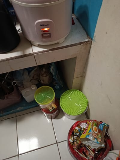 The image depicts a cluttered corner of a kitchen counter in Depok, Indonesia. A pink rice cooker with a lit orange indicator light is visible on the top shelf. Below, the shelves are filled with various items, including plastic containers, bags of groceries, and a ceramic teapot. On the floor, a red plastic bag overflows with instant noodle packets and other packaged snacks. The surrounding surfaces are tiled and appear somewhat aged. The scene suggests a domestic environment with everyday items accumulated.
