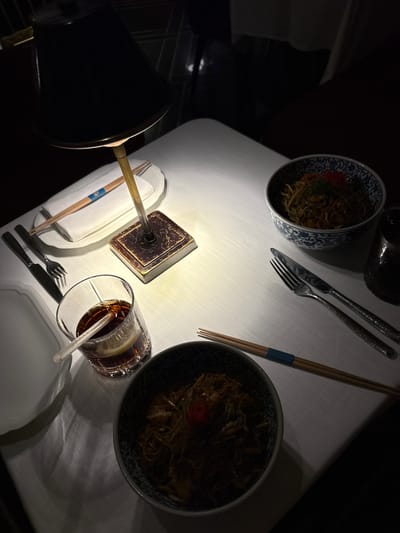 A table setting in Doha, Qatar, features two bowls of noodles, one with blue and white patterned edges and a red garnish, the other dark and with a similar garnish, placed on a white tablecloth. Silverware sets accompany each bowl, along with chopsticks, one set bundled with a blue band. A glass of dark liquid with a lemon slice and straw sits near one setting, adjacent to a small lamp with a square base and a conical shade. A folded white napkin is positioned near the lamp. The lighting casts shadows, suggesting an intimate dining atmosphere.