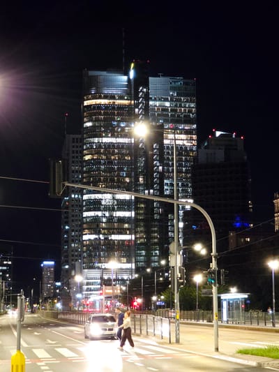 The image captures a nighttime scene on a city street in Warsaw, Poland. In the background, a large, modern skyscraper dominates the frame, its windows illuminated, creating a grid of light against the dark sky. To the right of the skyscraper, another building with dark windows and red lights on its upper section is visible. In the foreground, a street with multiple lanes is depicted, with white pedestrian crossings marked on the asphalt. Several cars are present on the road. Two pedestrians, a man and a woman, are crossing the street on a pedestrian crossing. The woman is walking, while the man appears to be gesturing. Streetlights and traffic lights illuminate the scene, casting a glow on the road and surrounding structures. A bus stop shelter with bright lighting is situated on the side of the road. The overall atmosphere is urban and active, characteristic of a city at night.