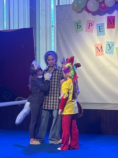 This is a vertical, full-shot, eye-level media file of what appears to be a children's theatrical performance. Three young people are on a stage, which is decorated with balloons on the left and colorful paper squares with Cyrillic letters on the white backdrop. The letters visible spell out "ВРЕМЯ" (VREMYA), meaning "time" in Russian.

The person in the foreground, on the bottom, is dressed in a dark outfit with a white furry tail and ears, suggesting a costume. They are looking up towards the person in the middle. The middle person is wearing a plaid shirt over a grey t-shirt and appears to be looking down at the person in front of them. This individual has on jeans and is leaning forward. The person at the back is wearing a bright yellow shirt, red leggings, and a colorful, spiky wig, indicating a character costume. They are also leaning forward, engaged with the others. The activity appears to be a dramatic scene from a play or performance, with the individuals interacting closely.

The lighting suggests an indoor setting, possibly a school or community theater. The blue floor of the stage is visible. The overall mood seems focused and theatrical, with the costumes adding a playful element. The time of day is not discernible, nor is the weather. The location context of Veliky Novgorod, Russia, is noted.