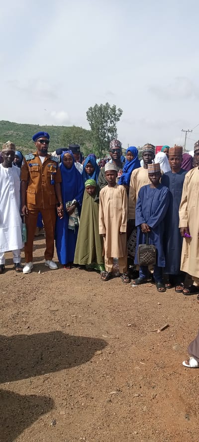 A group of people stand together in an outdoor area in Naraguta Hausawa, Nigeria. The ground is dirt and gravel. In the background, there is a hill with sparse vegetation and a partly cloudy sky. The group includes men, women, and children, most of whom are wearing traditional Islamic clothing. One man stands out due to his formal brown uniform with a blue beret and sunglasses. He holds hands with a woman in a blue headscarf. Several people are wearing traditional Islamic caps. A boy in a blue robe carries a small black patterned bag.
