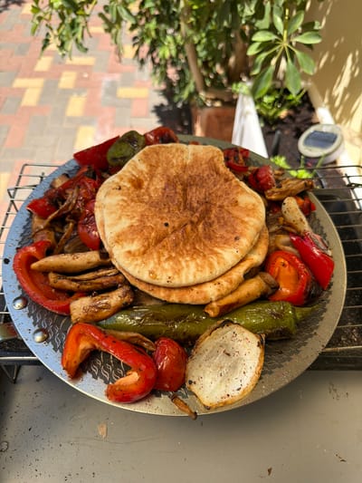 This is a top-down, slightly angled shot of a grilling basket filled with grilled vegetables and flatbread. The vegetables include red bell peppers, green peppers, cherry tomatoes, and what appear to be king oyster mushrooms and onions, all showing signs of charring from the grill. The flatbread, possibly pita, is golden brown and stacked in the center of the basket. The grilling basket sits on top of a metal grill grate.
In the background, to the left, there is greenery from a plant, and some soil. A small solar-powered garden light is visible in the soil. The ground is paved with interlocking pavers in shades of beige, red, and yellow. The lighting suggests it is daytime, likely sunny, with shadows cast by the greenery. The overall impression is a casual outdoor meal preparation, possibly in a backyard or garden setting in Even Yehuda, Israel. There is no visible text in the image.
