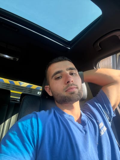 A young man with short dark hair and a beard is sitting in the driver's seat of a car, looking at the camera. He is wearing a blue t-shirt and has his right arm resting behind his head. The car's sunroof is open, revealing a clear blue sky. The interior of the car is visible, including the black leather seats and a section of the dashboard. In the background, there is a yellow and blue striped barrier, possibly from a construction site or road closure. The lighting suggests it is daytime, with sunlight casting shadows on the man and the car's interior. The image does not provide any specific details to locate it within the city of Gabrovo, Bulgaria.