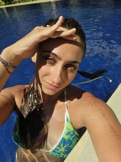 A female individual is partially submerged in a swimming pool in Pecatu, Indonesia, photographed from a high angle looking down. She appears to be a young adult with dark, wet hair, some of which is adorned with decorative beads and feathers on the left side. Her face is wet, with prominent green-hazel eyes, and she is looking directly at the camera. Her right hand is raised, palm flat against her forehead, shading her eyes from direct sunlight, which casts a shadow across her upper face. She is wearing a green and blue patterned bikini top. A thin, multi-colored beaded necklace is visible around her neck. Her left arm is visible, showing a grey woven bracelet and a red and black cord bracelet on her wrist. The left shoulder features a tattoo resembling sun rays, and a vertical geometric/symbolic tattoo extends down her left upper arm. The water in the pool is clear and blue, exhibiting ripples and reflections of sunlight. A light-colored pool edge is visible to the right.