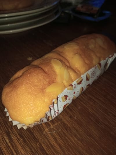 A golden-brown, almond-topped loaf cake, enclosed within a white paper liner featuring repetitive cake slice and spiral motifs, rests on a dark wooden surface in Khajuripada, India. The cake's upper surface displays a textured finish with numerous slivered almonds. In the blurred background, a stack of at least three light-colored plates is visible to the upper left. To the upper right, a dark, indistinct object, possibly a container with a blue label, is present. No individuals, animals, or active interactions are depicted within the scene.