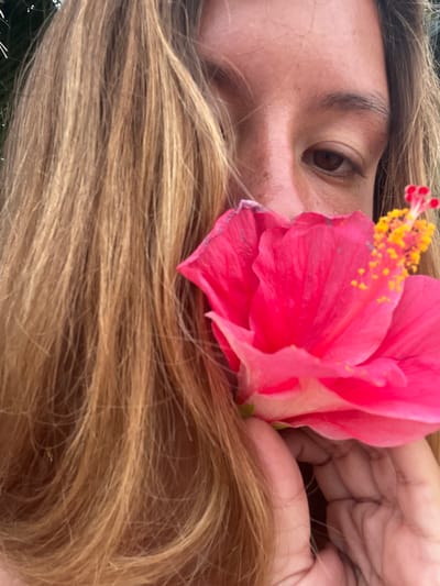 This is a close-up, high-angle medium shot of a person's face and a hibiscus flower in General Luna, Philippines. The person, seen from the eye down, has light brown hair that is partially covering their face and is looking towards the camera. Their skin has freckles, and they are wearing a pink shirt. The hibiscus flower is bright pink and is held close to their face by a hand that is also visible in the shot. The background appears to be natural, possibly with greenery, suggesting an outdoor setting.