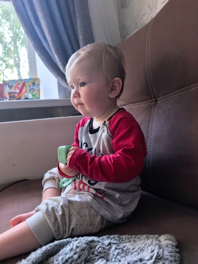 A young Caucasian child with blonde hair sits on a brown sofa. The child is wearing a grey long-sleeved shirt with red sleeves and grey pants. The child is holding a light green toy in their hands. The child is looking out a window with blue curtains. The window sill has a toy box on it. The background is a brown sofa. The image appears to have been taken indoors. There is no information available about the location in the city of Sterlitamak, Russia.
