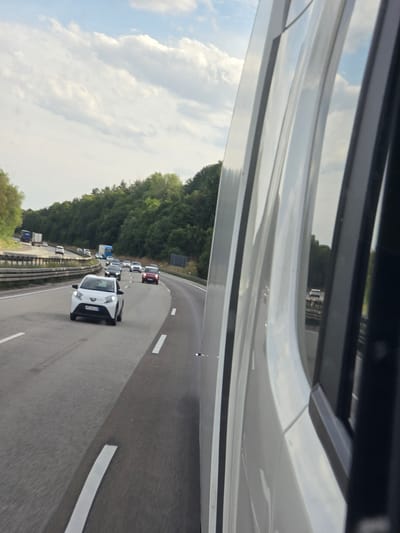 The image captures a view from inside a vehicle on a highway, likely traveling east or southeast. The foreground is dominated by the white, curved paneling of the vehicle's interior, with a dark vertical element on the right side. The highway itself is visible in the midground, with multiple lanes of traffic. A white hatchback is prominently featured in the left lane, followed by other vehicles. The highway is bordered by a metal guardrail on the left and a dense line of green trees on the right, characteristic of a rural or semi-rural setting. The sky above is partly cloudy with white clouds against a pale blue backdrop. The overall scene suggests a sunny day on a well-trafficked German autobahn, possibly near Wörsdorf, Germany, given the presence of German license plates on some vehicles.