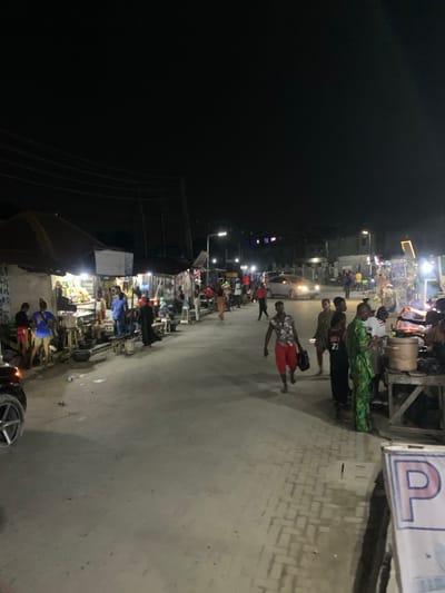The image depicts a nighttime street scene in Iranla, Nigeria, characterized by artificial illumination. The environment consists of a paved road with an adjacent sidewalk on the right, bordered by multiple makeshift market stalls and permanent structures on both sides. Overhead utility lines are visible against the dark sky.
Approximately 20-30 individuals are present. On the left, several people congregate near stalls displaying various goods under bright white lights. A dark SUV is partially visible in the foreground on the left. In the mid-ground, a person wearing a floral shirt and red shorts walks towards the viewer, holding a dark bag. Other individuals are walking along the road and congregating near stalls on the right side. A light-colored sedan is visible in the distance, heading away from the viewer.
On the right side, multiple stalls are active, with individuals standing, interacting, and preparing items. One stall in the foreground features a large metal pot on a wooden stand. A partially visible blue and white sign with the letter "P" is in the lower right corner. The ground shows some scattered debris.