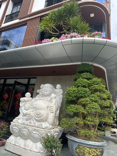 This image captures the exterior of a modern, multi-story building located in Hòa Cường Ward, Vietnam, under daylight conditions. The building exhibits a contemporary design with a prominent, curved, silver-paneled overhang extending over the ground floor entrance. Above this overhang, the second story features large glass windows flanked by brown vertical slats and a balcony adorned with several large, spiky-leaved plants and numerous smaller flowering plants displaying pink, purple, and yellow blossoms.

On the ground level, to the left of the main entrance and directly beneath the overhang, a large, white marble statue of a seated Budai (Laughing Buddha) is present. This central figure is depicted with a smiling expression and is surrounded by five smaller child figures. The statue rests on a multi-tiered lotus-shaped base, also carved from white marble. To the right of the Budai statue, a large, meticulously shaped bonsai tree with dense green foliage stands within a decorative grey and gold pot.

The building's ground floor entrance consists of dark-framed glass doors, through which red decorative items featuring golden Chinese characters are visible. A smaller potted plant with reddish foliage is positioned near the base of the Budai statue, and another pot with red flowers is partially visible further to the left. The immediate ground area appears to be a light-colored paved surface. A motor scooter is partially visible on the far right side of the frame, beyond the large bonsai tree.