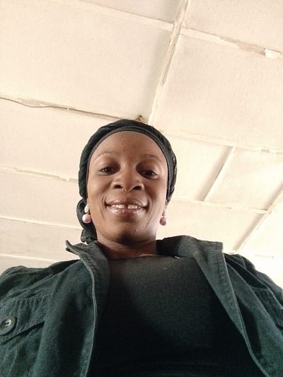A single adult female, appearing to be of African descent, is depicted from a low-angle perspective, facing the camera with a broad smile revealing a distinct gap between her upper front teeth. She wears a dark-colored, possibly black, textured headwrap, and light pink spherical drop earrings with decorative tops. Her attire includes a dark-colored, likely black or dark grey, collared jacket or shirt with visible buttons on the left lapel, over a plain dark top. The background consists of a light-colored (off-white or light beige) paneled ceiling with visible grid lines, seams, and minor discoloration or wear. A thin, dark wire runs horizontally across the upper left section of the ceiling. The scene takes place indoors in Barnawa, Nigeria.