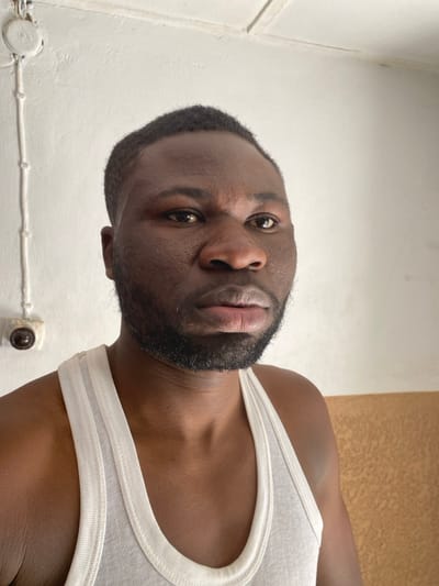 The depicted location is identified as Udu, Nigeria. The image shows a close-up, head-and-shoulders shot of an adult male with dark skin, dark short hair, and a short beard and mustache. He is wearing a white ribbed tank top and looks directly at the viewer with a neutral expression, his head slightly tilted. The background consists of a plain white wall. On the left side of the wall, a white electrical conduit runs vertically, secured by clips, with a round, dark brown electrical component, possibly a switch, attached to it at shoulder height. The lower right portion of the wall indicates a transition to a tan or light brown colored section.