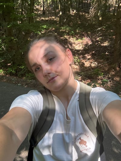 A person is taking a selfie outdoors, with a forested area in the background. The person is wearing a white t-shirt with a graphic on it and a green backpack. The graphic on the t-shirt includes a silhouette of a horse and text that reads "Memphis Tennessee usa". The image appears to be taken during the day, with sunlight filtering through the trees and casting shadows. The person's face is partially visible, and they have a slight smile. The setting suggests a walk or hike in a natural environment. The presence of the backpack implies they may be traveling or engaged in an outdoor activity. The overall mood is relaxed and casual. There are no specific cues to indicate the exact time of day beyond it being daytime, or the specific weather conditions other than it appears to be a sunny day. No specific details about Au in der Hallertau, Germany are visible in the image.