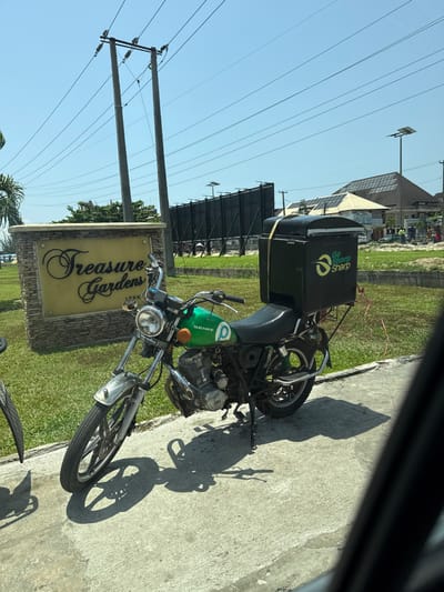 The image depicts an outdoor daytime scene at Treasure Gardens IEKKU, located within Alaguntan, Nigeria. A green "SENKE" brand motorcycle is centrally positioned, parked on a light gray concrete surface, supported by a kickstand. The motorcycle features a large, black, rectangular delivery box affixed to its rear rack, secured by a yellow strap. The box displays a white and green "Go Sharp Sharp" logo. To the left and slightly behind the motorcycle, a stone-faced sign monument with a golden plaque reads "Treasure Gardens" in cursive script, with "IEKKU" inscribed in smaller text below it. The ground surrounding the sign and extending into the background is covered with green grass. Overhead, against a clear blue sky, multiple wooden utility poles with electrical lines are visible. In the background, dark, tall billboard-like structures, several solar-powered streetlights, and a building with a multi-paneled dark roof are present. Indistinct human figures are visible in the far background near the building, possibly engaged in activity. A partial view of a vehicle's interior frame is visible on the left and right edges of the foreground.