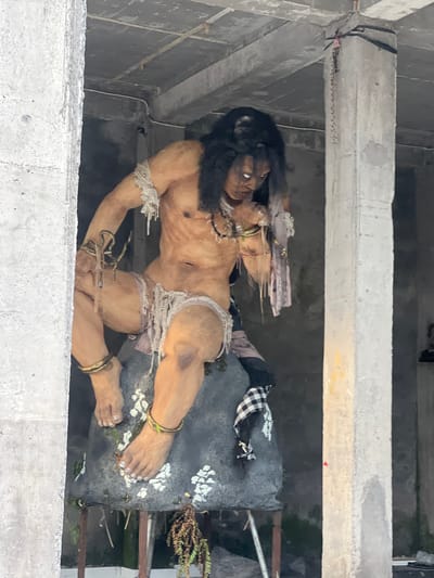The image shows a large, detailed statue of a dark-haired male figure lying on a gray, textured surface. The statue appears to be made of a material resembling clay or resin and is predominantly tan, with darker accents around the hair and facial features. The figure is partially draped in rough fabric, including a white and black checkered cloth.
The statue is positioned within a gray, concrete structure, possibly the interior of a building or enclosure under construction. The view is from above, looking down into the space where the statue is placed. The concrete structure consists of horizontal beams or ledges. Based on context, this is likely a religious statue in Celuk, Indonesia.