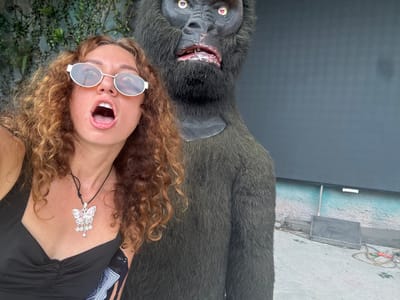 A woman with curly brown hair and sunglasses is taking a selfie in front of a large, furry ape statue. The woman is wearing a black top and a butterfly pendant necklace. The ape statue is dark gray and has realistic features, including fur, eyes, and a mouth with visible teeth. The background appears to be an outdoor setting with some greenery and a dark wall. The ape statue's left arm is extended, and the woman's left arm is visible as she holds the phone to take the picture. The image is taken from a medium shot, with the woman and the ape statue occupying most of the frame.