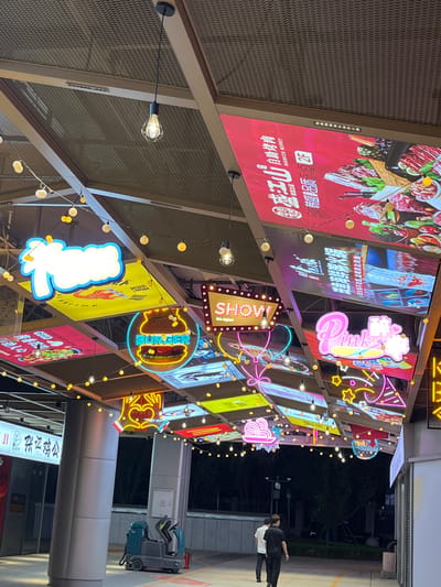 The image shows an outdoor food court area with various neon signs and banners hanging from a metallic ceiling structure. The signs advertise different food options, including a burger neon sign and banners featuring images of meat and seafood. String lights with round bulbs are also visible, illuminating the area.

In the foreground, two men are walking away from the camera. To the left, a cleaning machine is parked near a pillar. The overall environment appears to be a modern commercial area, likely a street or plaza within the Daxing District of China, as suggested by the signage and architectural style. The lighting indicates it is either dusk or night.