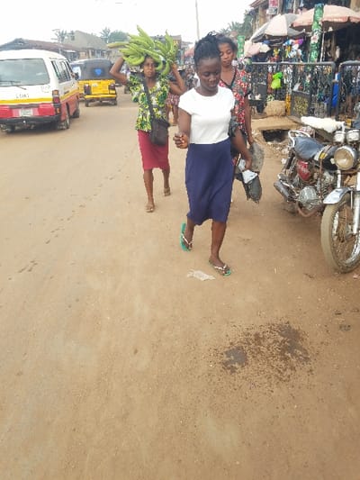 The image depicts a street scene in the city of Nnobi, Nigeria, characterized by an unpaved, light brown dirt road under an overcast sky. Three adult females are walking from left to right across the frame.
In the foreground, a woman wears a white short-sleeved top, a navy blue skirt, and green flip-flops, holding a black plastic bag in her right hand. Her gaze is directed downwards. Behind her, a second woman carries a large, prominent bunch of green plantains on her head. She is attired in a vibrant, patterned short-sleeved top, a red knee-length skirt, and sandals, with a black messenger bag slung over her left shoulder. Partially visible behind the second woman is a third female, wearing a patterned top.
The right side of the street features a row of market stalls or shops with awnings and various goods, fronted by a black metal barrier. Two motorcycles are parked vertically along the right edge of the road. In the left background, a white mini-bus with red and yellow markings is visible, followed by a yellow tricycle (keke napep). Utility poles and overhead wires are present in the distant background. The road surface in the immediate foreground exhibits a dark, damp patch and some scattered debris.