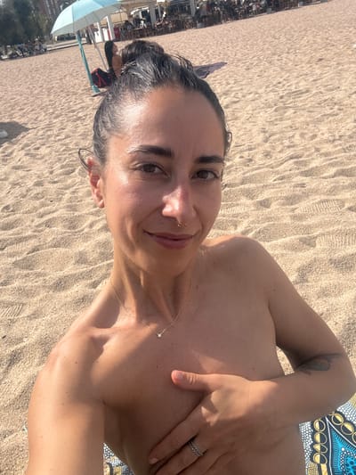 A woman is taking a selfie on a sandy beach in Badalona, Spain. She is topless and holding her left hand over her chest. She is wearing a gold necklace with a small diamond pendant and a ring on her left ring finger. The beach is populated with people in the background, some sitting at tables and others walking. There is a light blue beach umbrella in the background. The sand is light brown and appears to be wet in some areas. The sun is shining, casting shadows on the sand. The woman has her hair pulled back in a bun and is wearing a nose ring. She has a tattoo on her left arm.