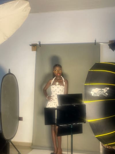 An individual stands centered in a studio setting, facing the camera. They are wearing a white halter dress with black polka dots, a necklace, and a bracelet on their left wrist. Their left hand is on their hip, and their right arm is bent with the hand resting on their hip. The background is a light grey seamless backdrop. To the left of the individual is a large, oval-shaped light reflector on a stand. To the right is a large, hexagonal softbox with yellow trim, displaying a logo with a world map and the letters "APCS". In front of the individual are two black monitors mounted on stands. The lighting appears to be studio lighting, with a large diffused light source at the top left of the frame and the softbox providing fill light. The floor is tiled in a light color. The overall impression is a professional or semi-professional photoshoot. There is no visible indication of the specific location within South Ropp, Nigeria.
