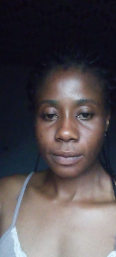 The image is a close-up selfie of a young Black woman. She has dark skin, braided hair, and is wearing a white tank top. She is looking down and to the side, with a neutral expression. The background is dark and out of focus, suggesting an indoor setting. There are no other people or objects visible in the frame, and no specific location in Kakau, Nigeria, can be identified from the image.